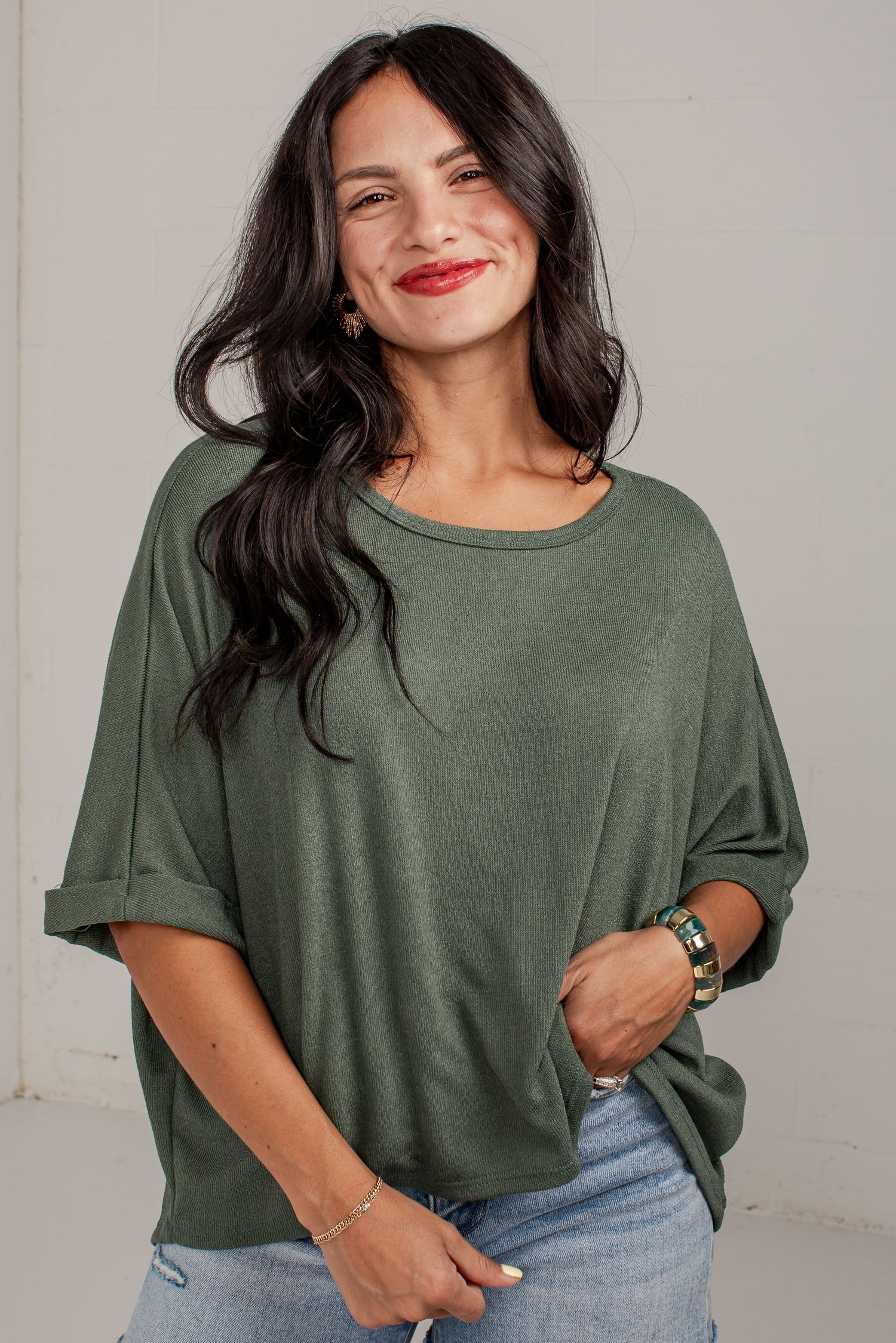 A woman modeling a loose-fitting, olive green short-sleeve top with rolled cuffs, paired with denim shorts, standing against a plain light gray wall.