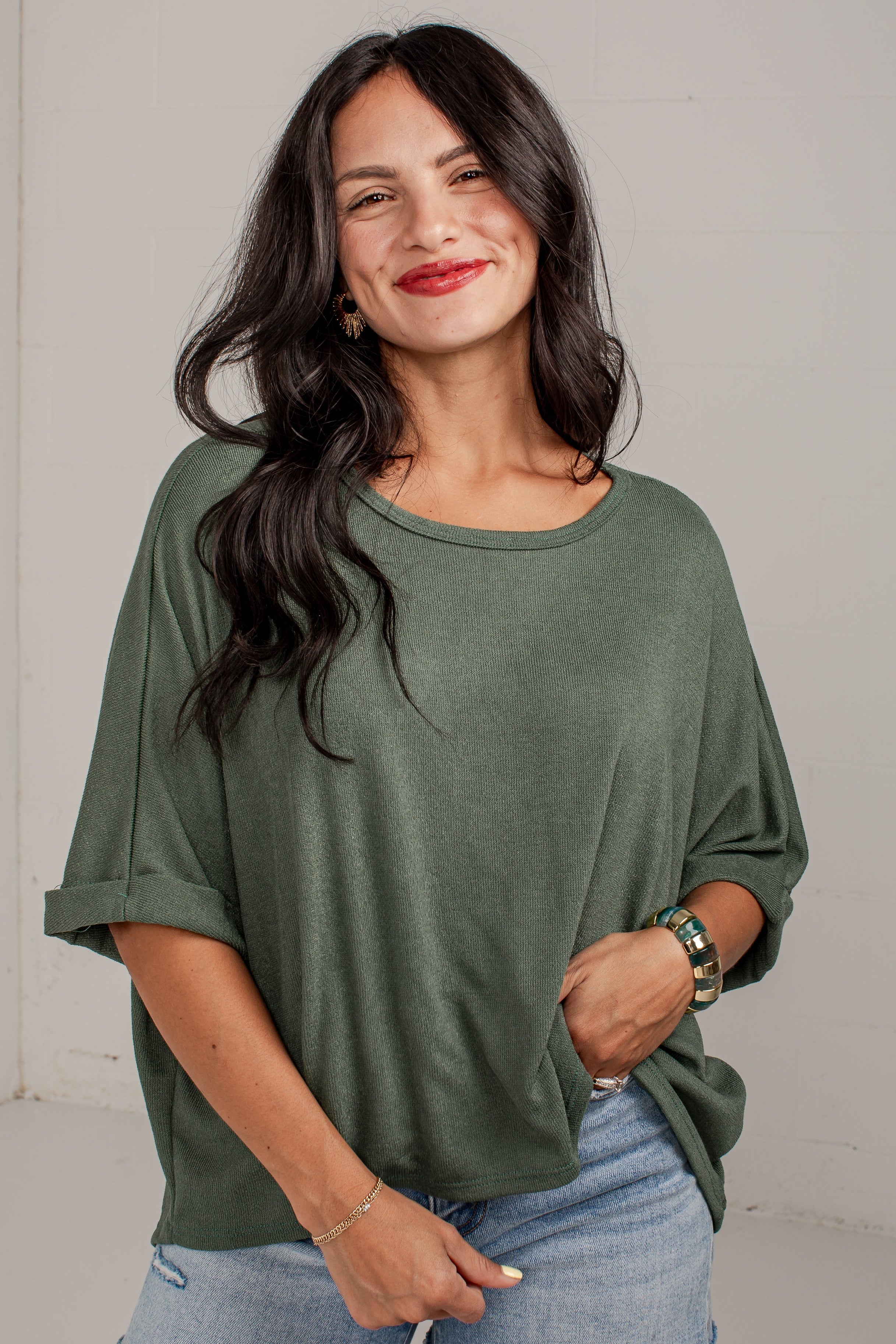 A woman modeling a loose-fitting, olive green short-sleeve top with rolled cuffs, paired with denim shorts, standing against a plain light gray wall.