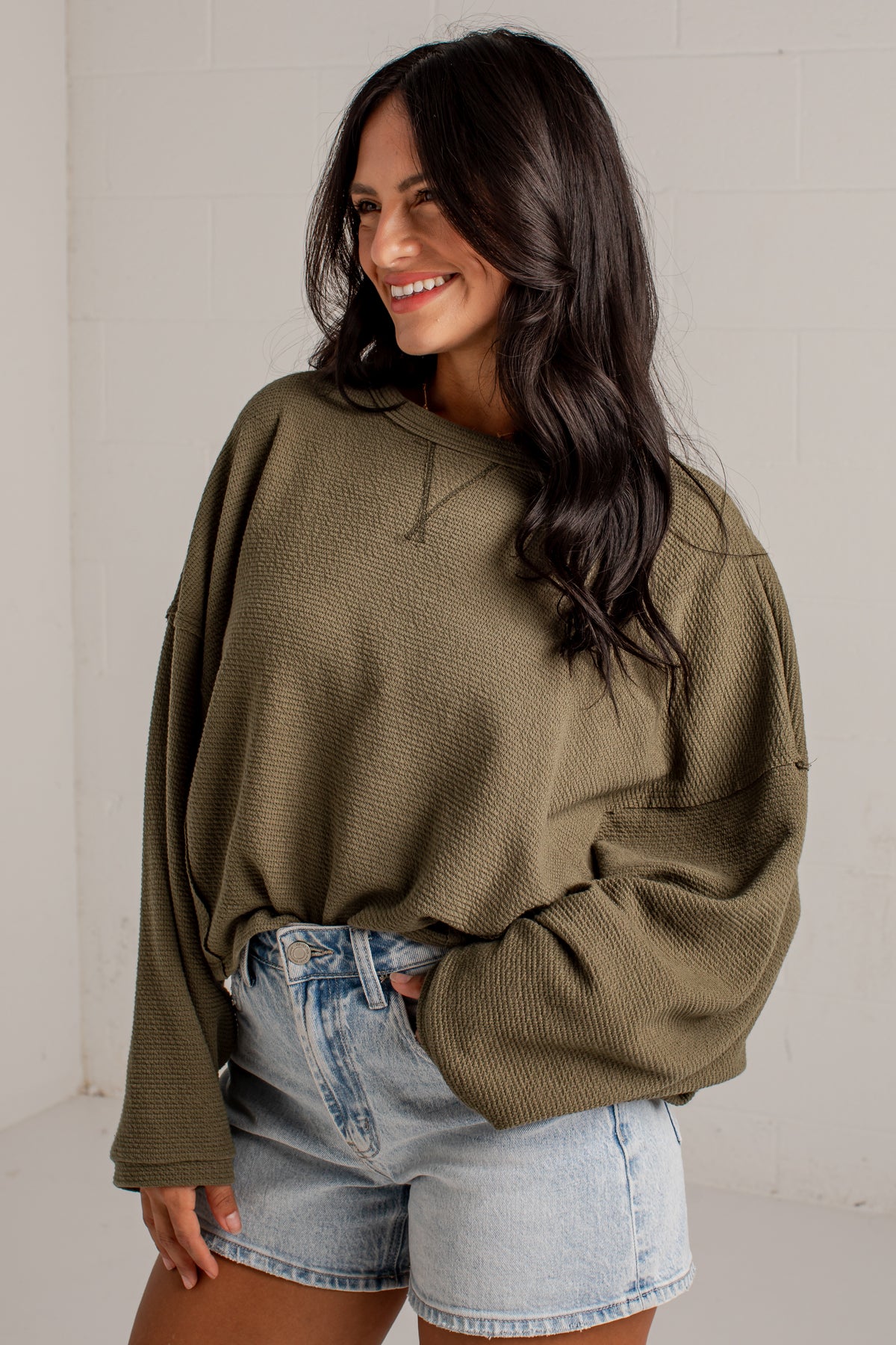 A woman wearing an oversized olive green waffle-knit long-sleeve sweatshirt and light-wash denim shorts, standing against a white brick wall.