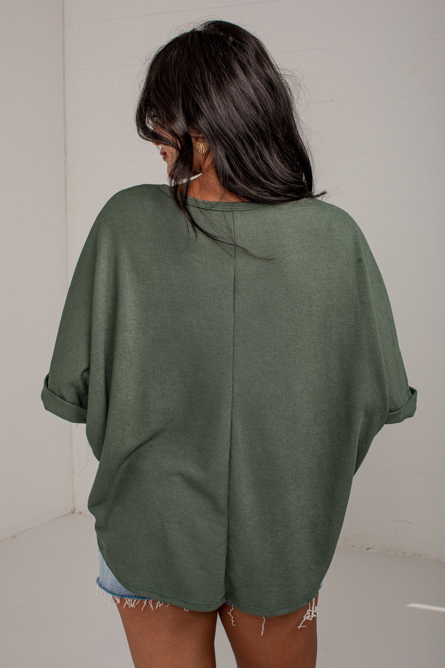 A woman viewed from behind wearing an oversized sage green knit top with rolled sleeves and a split back seam, paired with denim shorts.