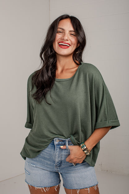 A woman smiling and wearing an olive green short-sleeve tunic top with light blue denim shorts, standing against a light gray wall.