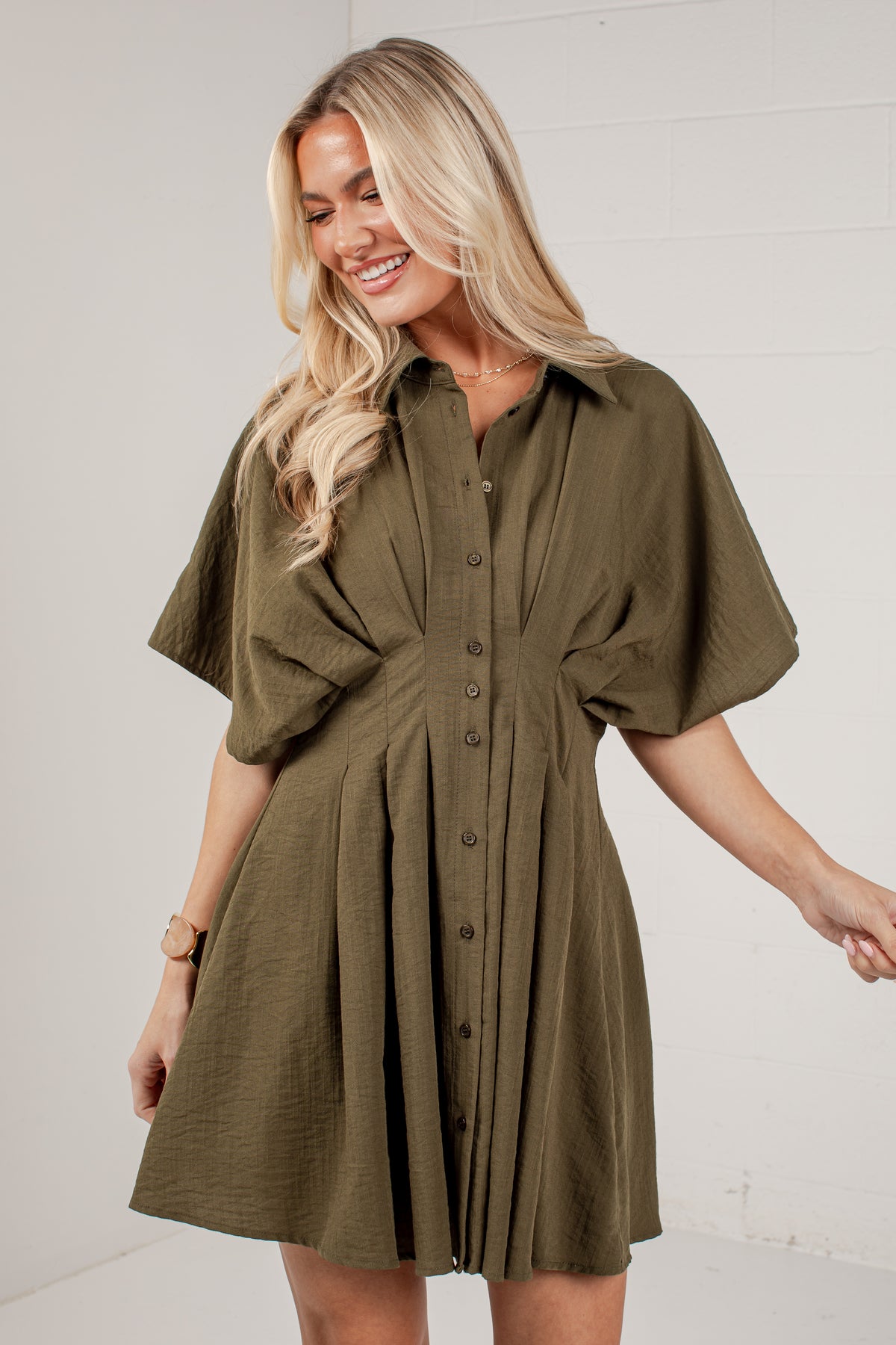 A woman wearing an olive green short-sleeve button-up shirt dress, standing against a white brick wall.