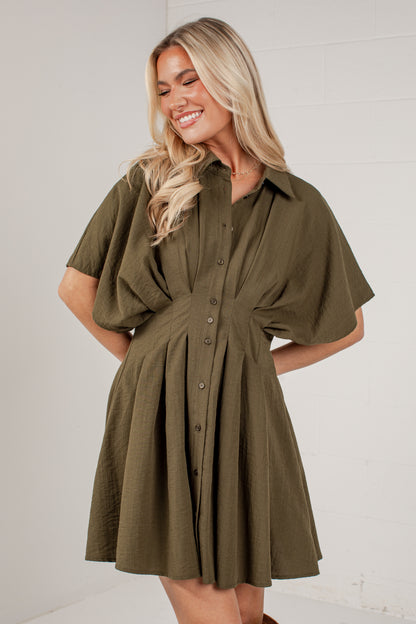 A woman wearing an olive green short-sleeve button-up dress with a gathered waist and flared skirt, standing against a white paneled wall.
