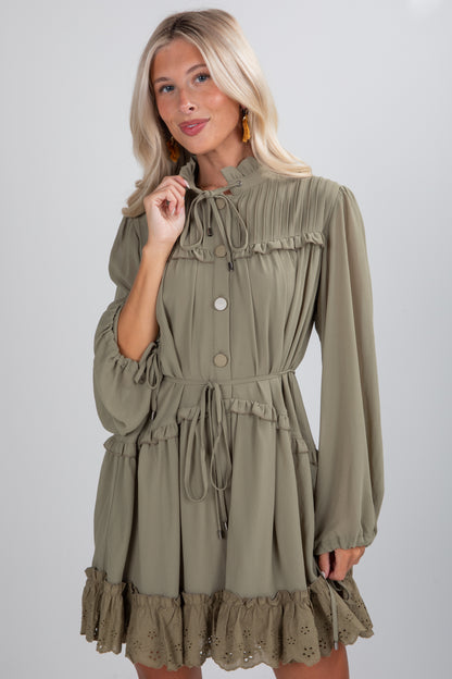 Woman wearing an olive green dress with long sleeves and ruffled details on a plain background