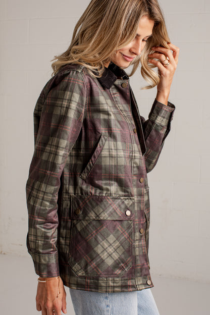 A woman wearing a long-sleeve plaid waxed jacket in muted green, brown, and burgundy tones, paired with light blue jeans, photographed against a light gray brick wall.