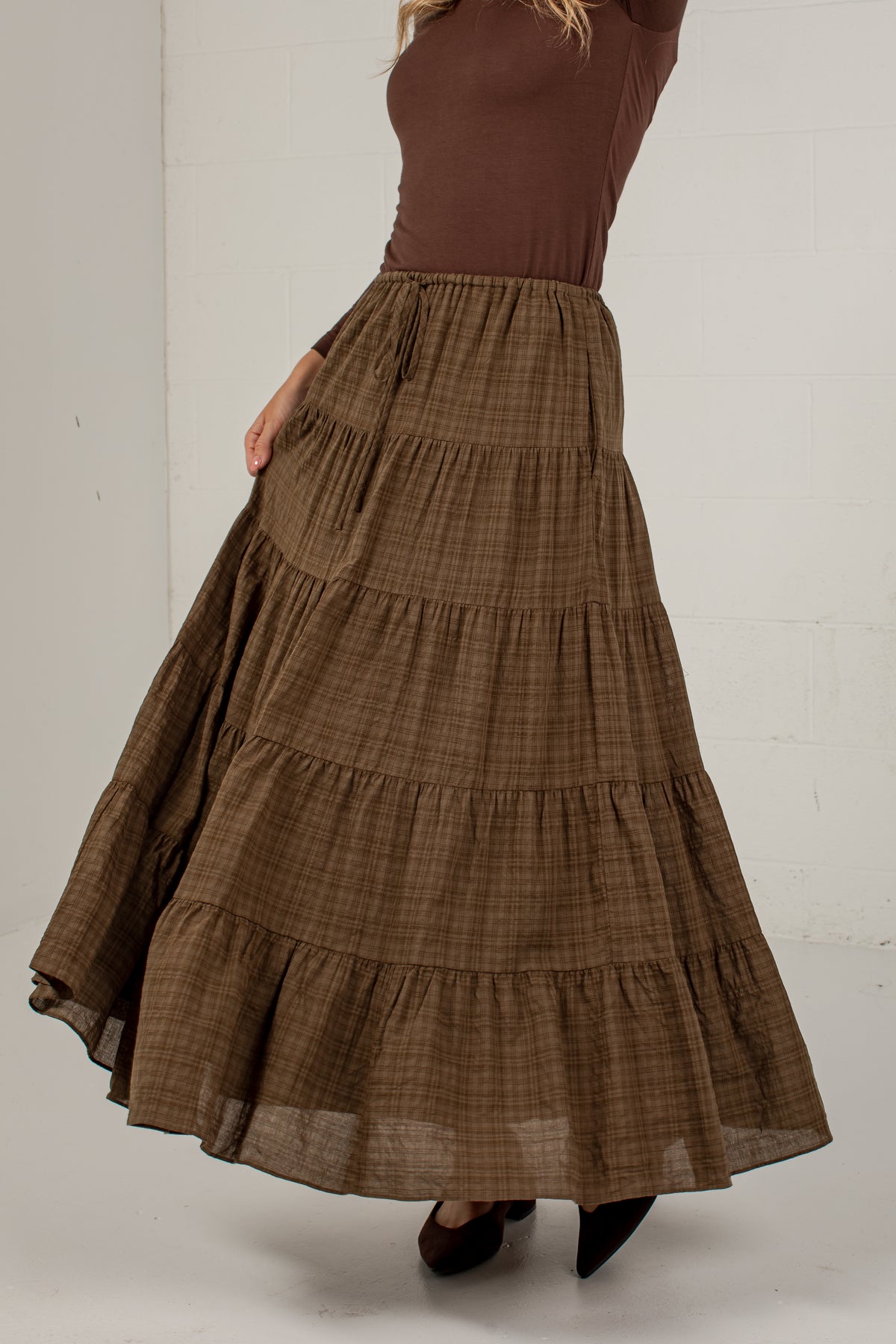 A woman wearing a brown tiered maxi skirt with a subtle plaid pattern, paired with a brown top and black pointed shoes, standing against a white wall.