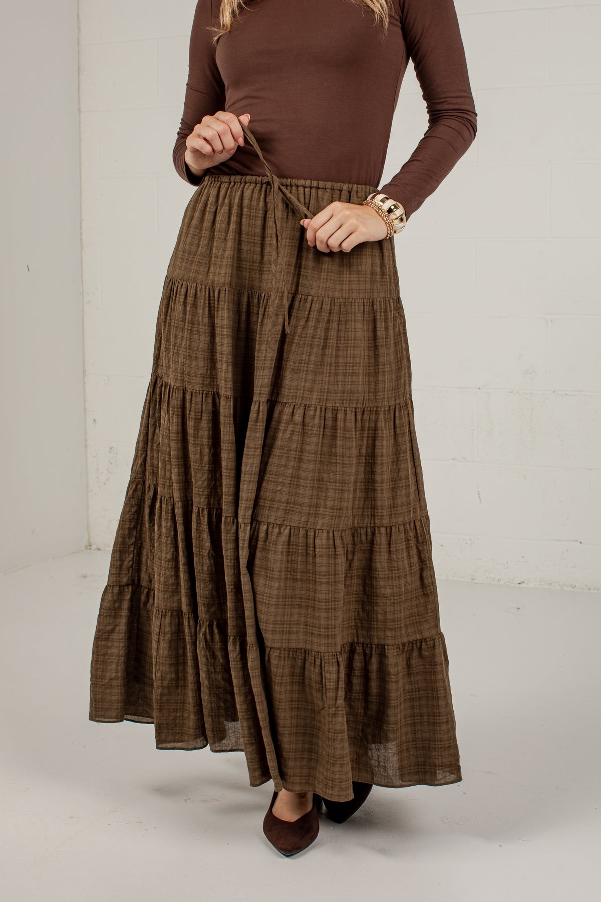 A woman wearing a brown tiered maxi skirt with a subtle plaid pattern, paired with a matching brown long-sleeve top and dark brown shoes, standing against a white wall.
