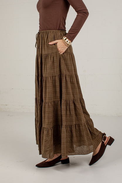 A model wearing a brown and beige plaid tiered maxi skirt with pockets, paired with a brown long-sleeve top and brown flat shoes, standing against a white brick wall.