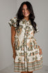 A woman modeling a short-sleeve button-front dress with a cream base and an intricate botanical pattern in olive green, rust orange, and black, set against a light gray brick wall.