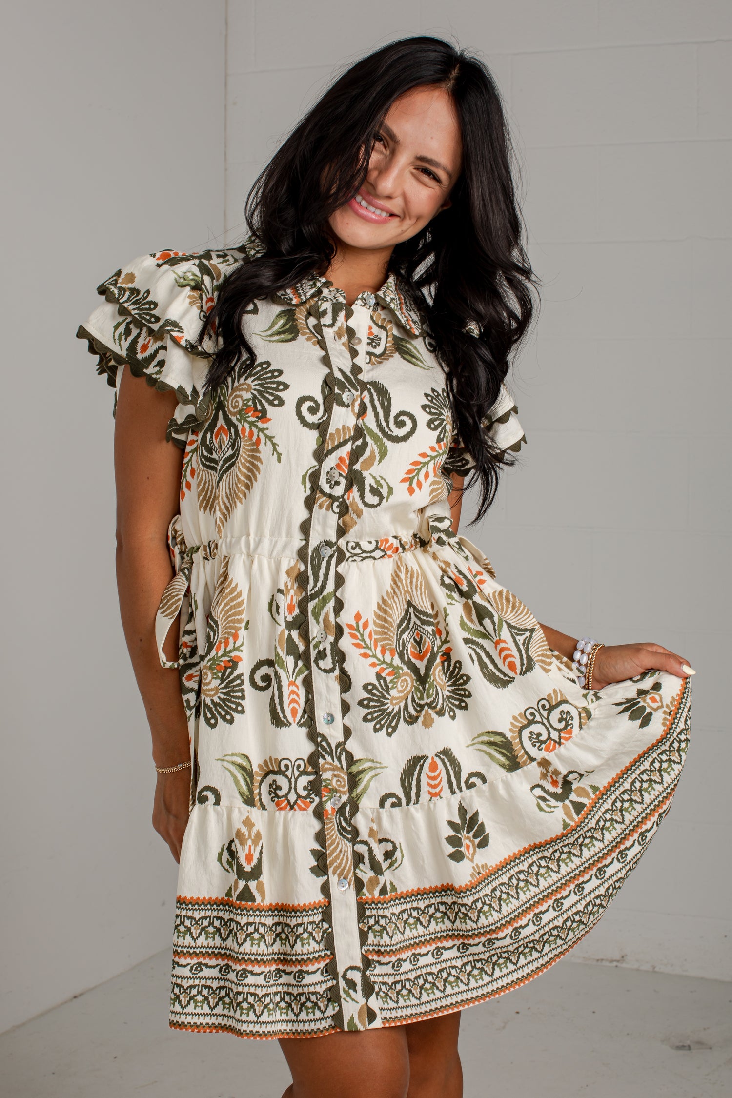 A woman modeling a short-sleeve bohemian-style mini dress with a cream base and green, brown, and orange floral patterns, set against a light gray wall.