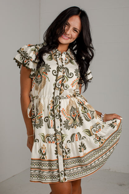 A woman modeling a short-sleeve bohemian-style mini dress with a cream base and green, brown, and orange floral patterns, set against a light gray wall.