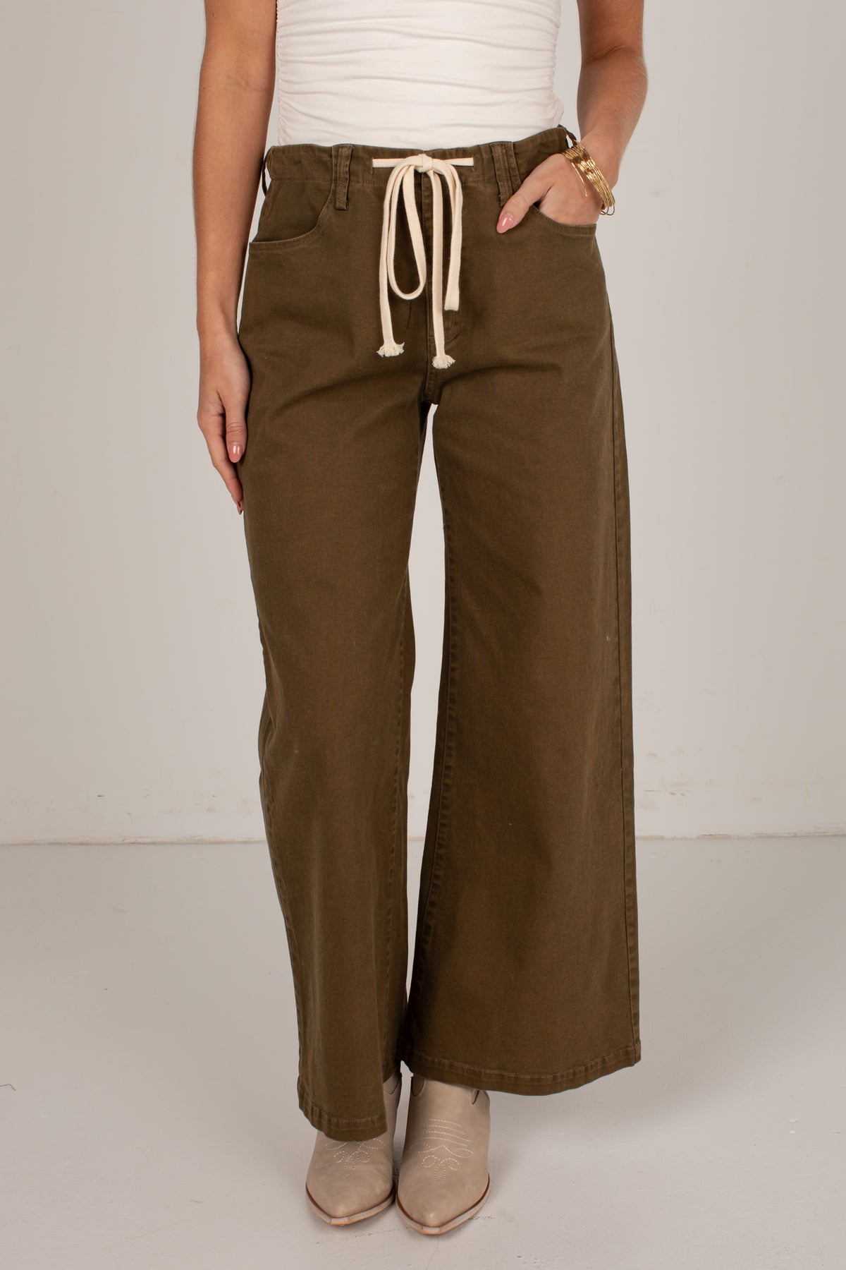 A person wearing olive brown wide-leg drawstring pants, styled with a white top and beige pointed-toe boots against a neutral studio background.