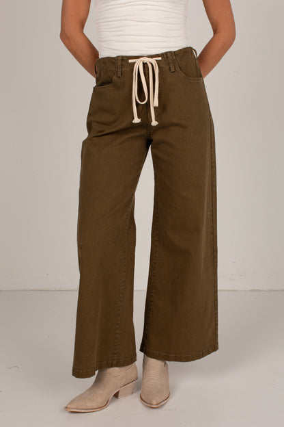 A person wearing wide-leg olive brown pants with a cream drawstring waist, paired with a white ribbed top and beige ankle boots, shown from the waist down against a plain light gray studio background.