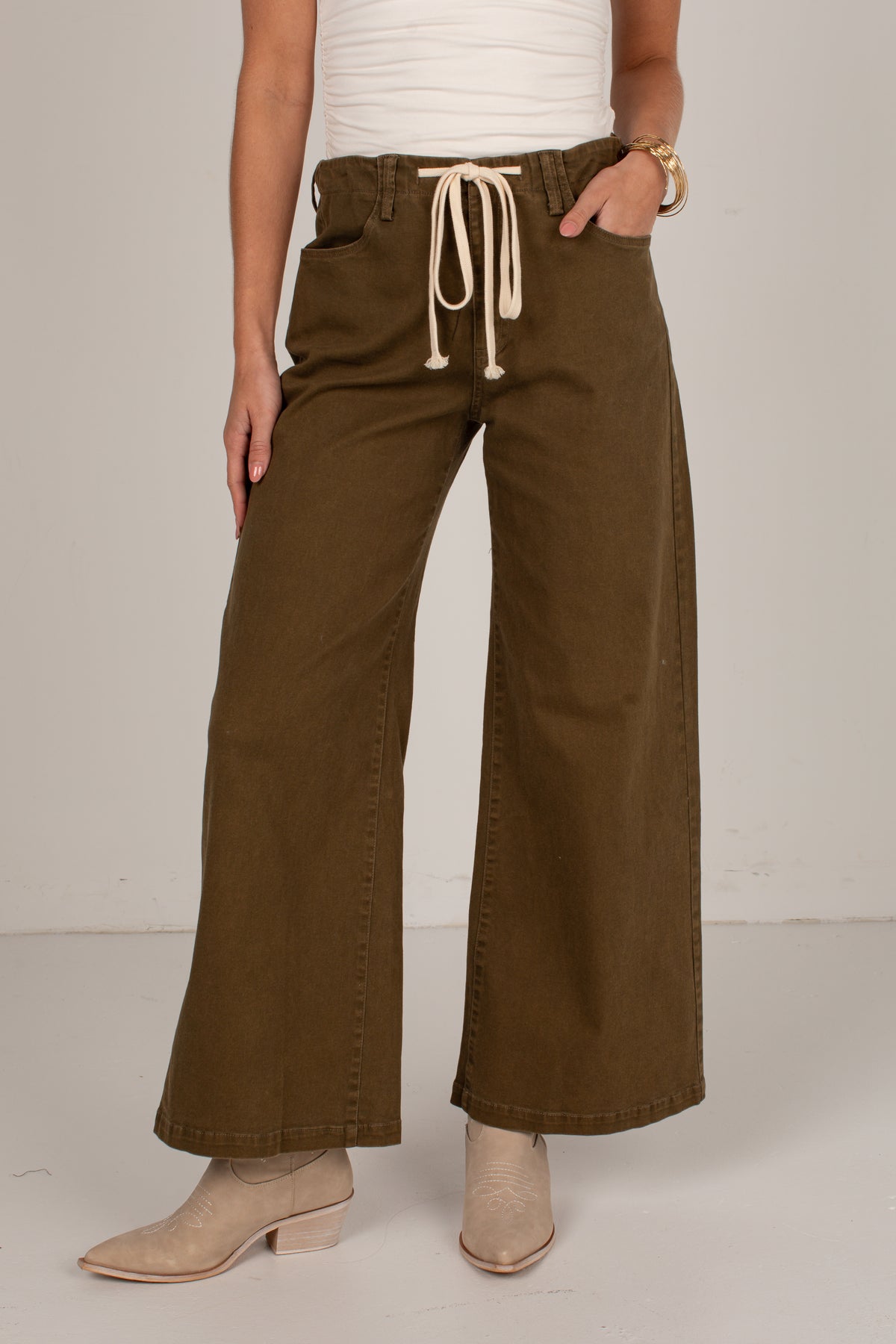 A person wearing olive brown wide-leg drawstring pants, a white ribbed top, and beige ankle boots, standing against a light gray studio background.