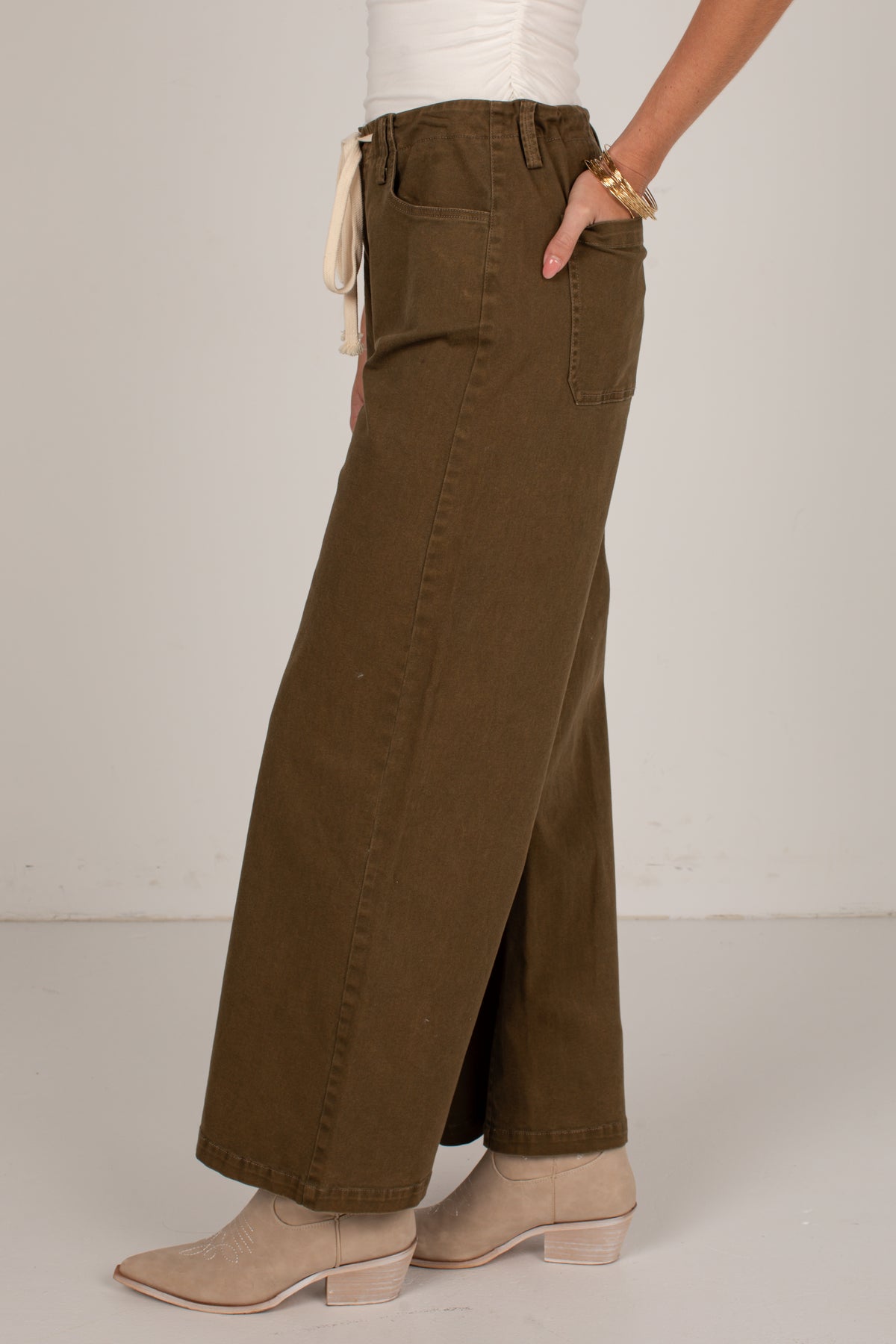 A person wearing high-waisted, wide-leg brown pants with a cream tie detail at the waist, paired with beige ankle boots against a plain off-white studio background.