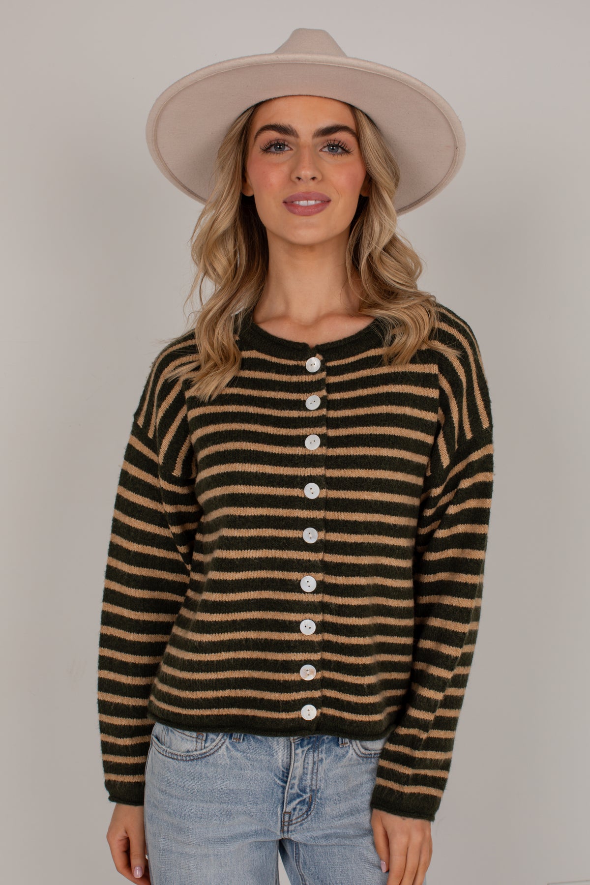 A woman wearing a dark olive green and tan striped knit cardigan sweater with white buttons, paired with light blue jeans and a beige wide-brim hat, against a plain light gray studio background.