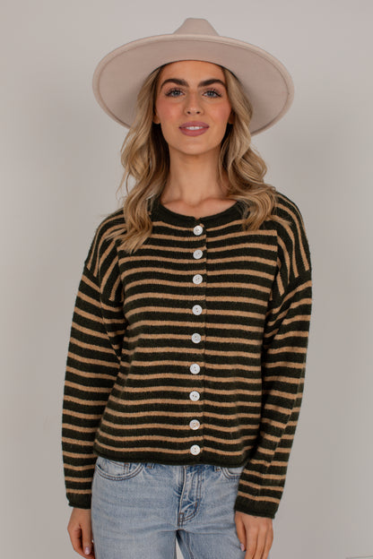A woman wearing a dark olive green and tan striped knit cardigan sweater with white buttons, paired with light blue jeans and a beige wide-brim hat, against a plain light gray studio background.