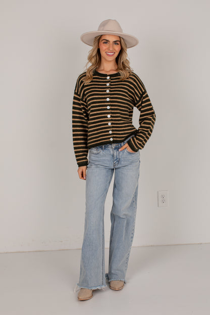 A woman modeling a brown and black striped button-front knit sweater, paired with light-wash flare jeans, beige flats, and a beige wide-brim hat, standing against a plain white wall.