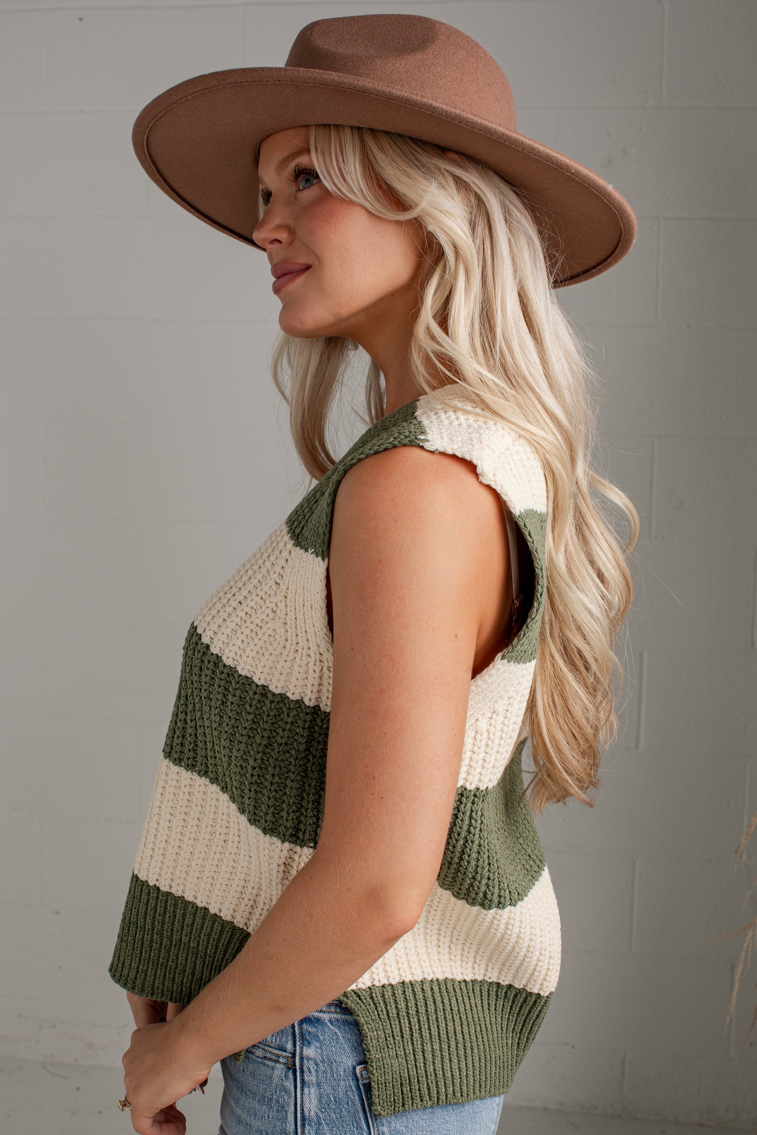 A woman wearing a brown wide-brim hat and a sleeveless knit vest with horizontal olive green and cream stripes, paired with blue jeans, photographed against a light gray brick wall.