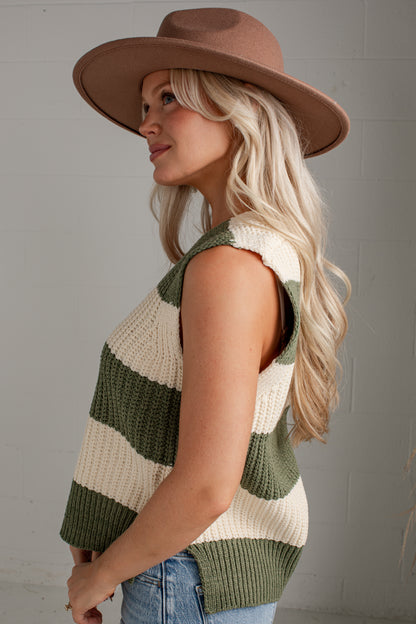 A woman wearing a brown wide-brim hat and a sleeveless knit vest with horizontal olive green and cream stripes, paired with blue jeans, photographed against a light gray brick wall.