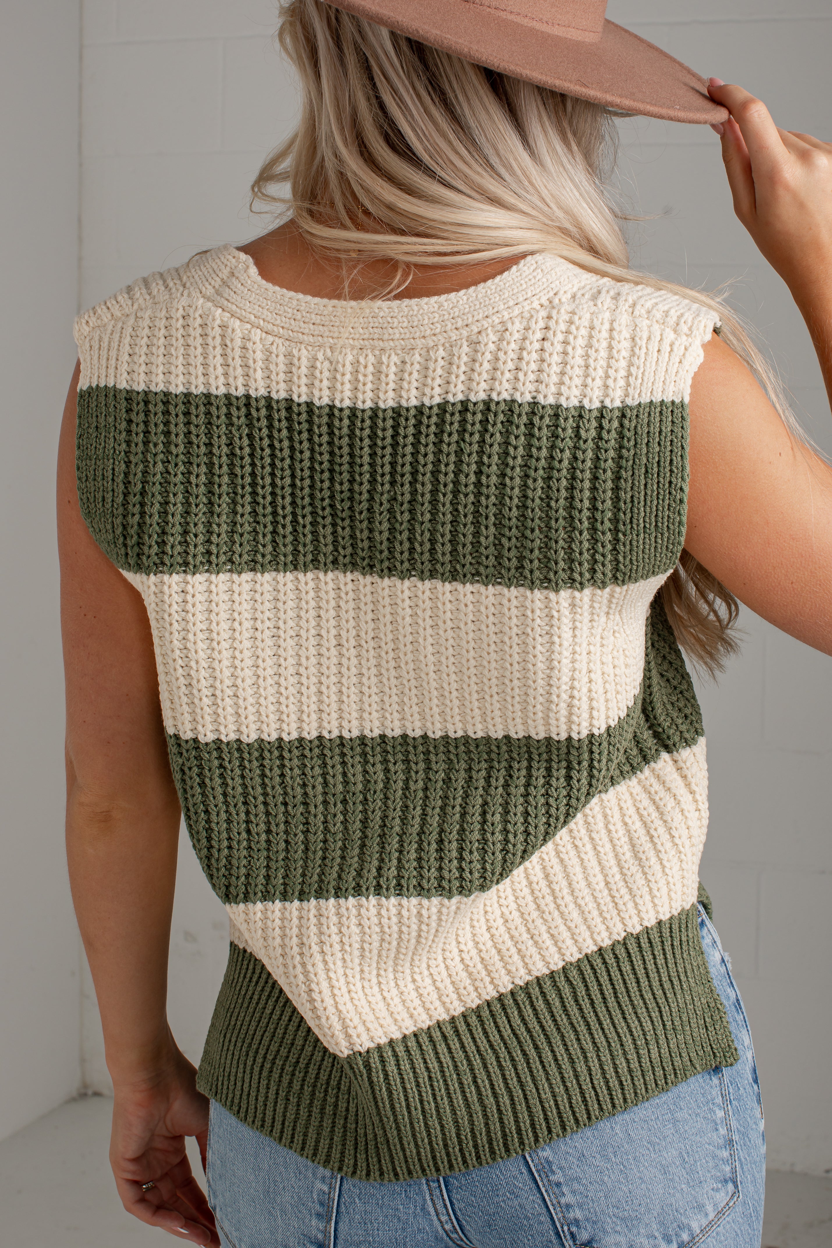 A woman shown from the back wearing a sleeveless ribbed knit sweater vest with horizontal stripes in olive green and cream, paired with light blue jeans and a dusty pink hat.
