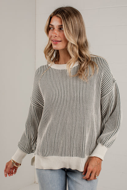 A woman wearing a cream and olive vertically striped knit sweater with balloon sleeves, paired with light blue jeans, standing against a white brick wall.