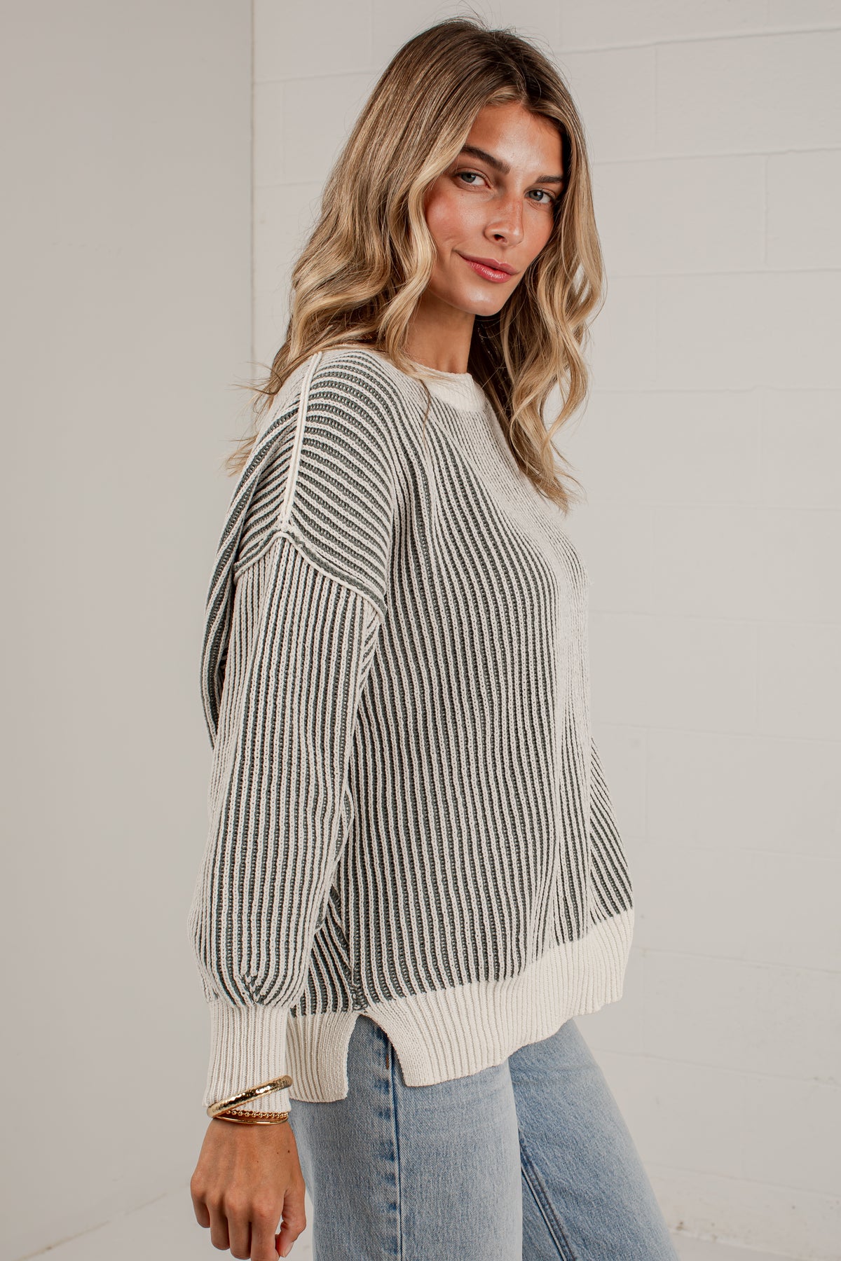 A woman wearing a gray and off-white vertically striped knit sweater with ribbed trim, paired with light blue jeans, standing against a white paneled wall.