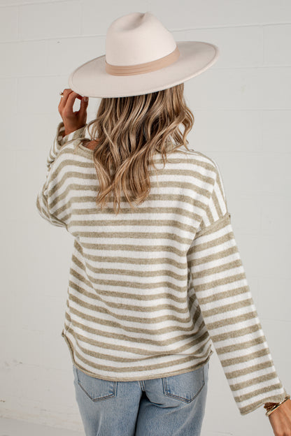 A woman shown from the back wearing a cream and tan striped knit sweater, a light beige wide-brim hat, and light blue jeans against a white paneled wall.