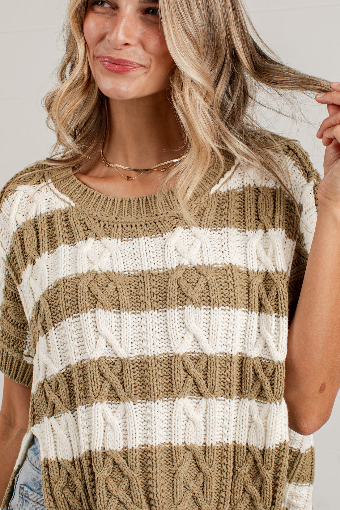 A woman wearing a short-sleeve knit sweater with horizontal stripes in taupe and off-white, featuring cable-knit patterns. The photo is a close-up crop of the upper body.