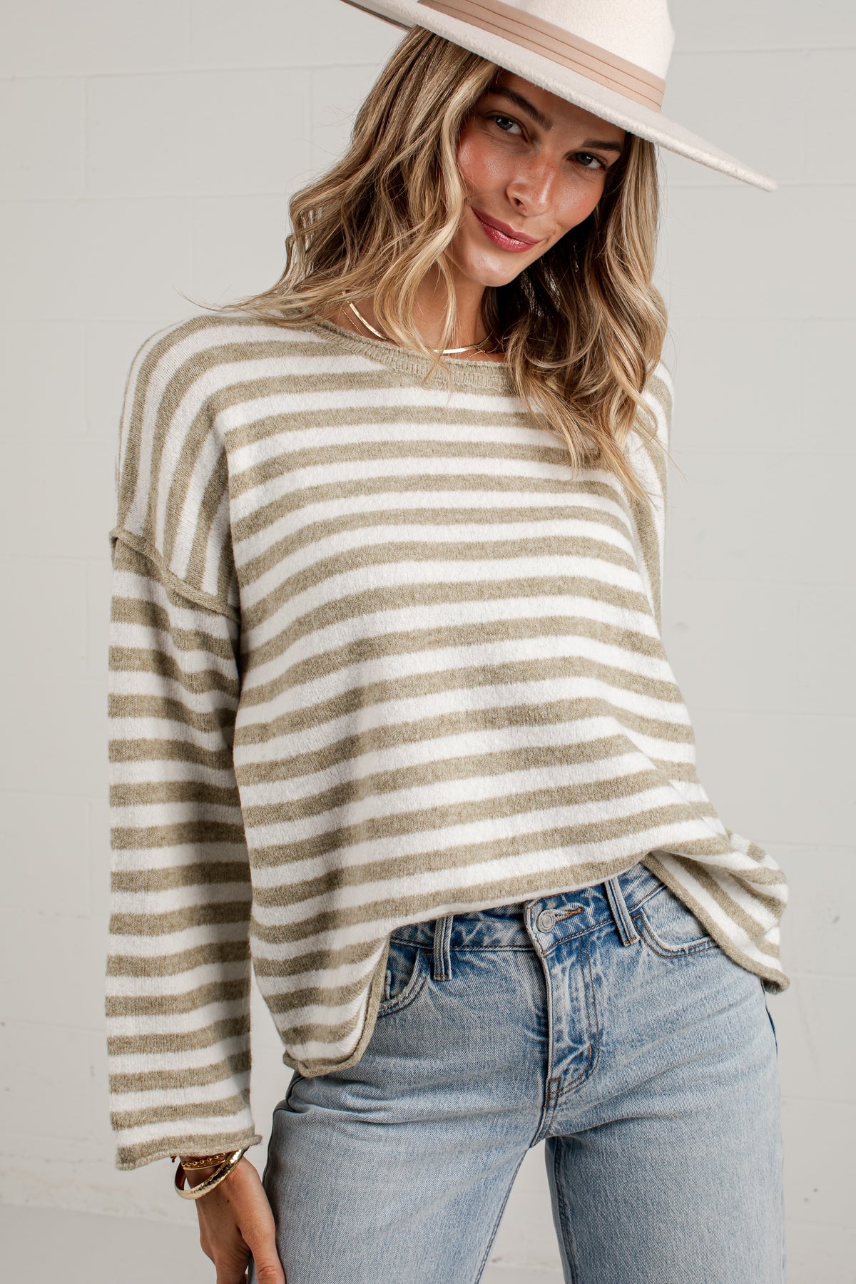 A woman wearing a beige and off-white horizontally striped knit sweater, light blue jeans, and a wide-brimmed beige hat, photographed against a light gray wall.
