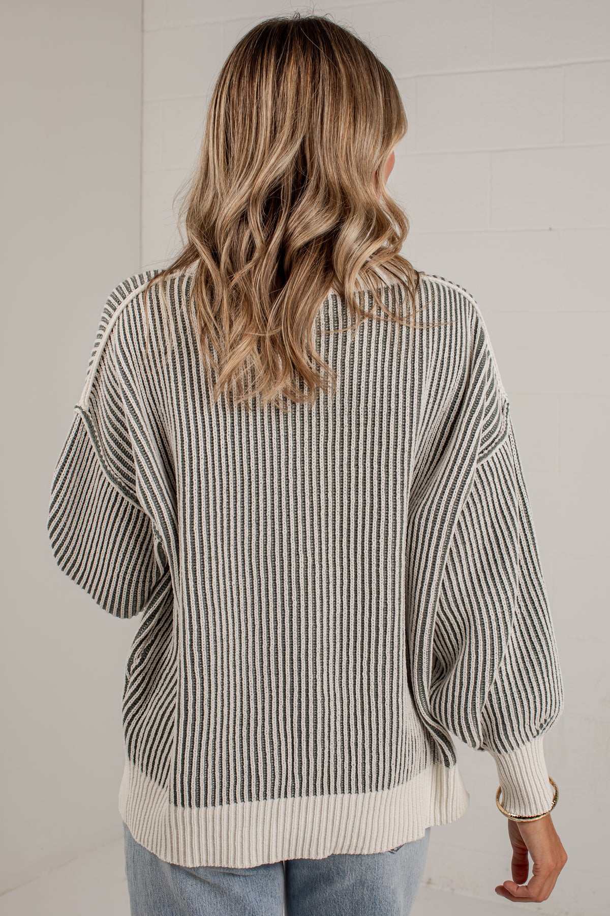 A woman viewed from behind wearing a cream and dark gray vertically striped knit sweater with balloon sleeves and ribbed trim, paired with light blue jeans against a white paneled wall.