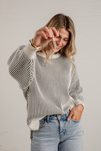 A woman wearing a cream and gray vertically striped knit sweater with a crew neck and dropped shoulders, paired with light-wash blue jeans, posing against a white paneled wall.