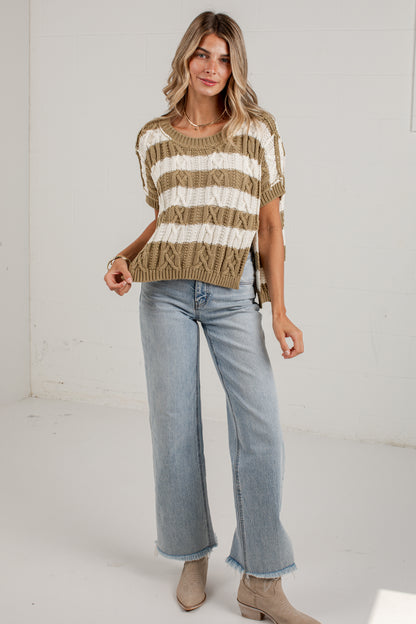 A woman modeling a short-sleeve knit sweater with horizontal beige and off-white stripes, paired with light-wash wide-leg jeans and beige ankle boots, standing against a white studio wall.