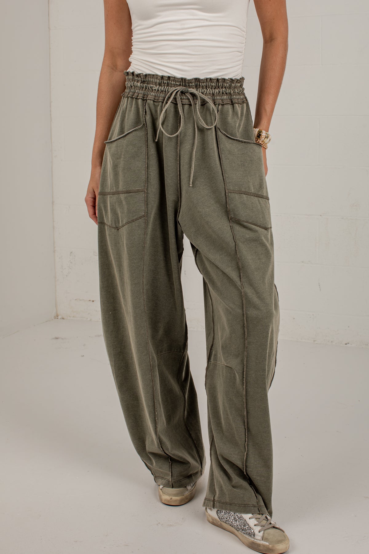 A person wearing loose, wide-leg olive green drawstring pants with large front patch pockets, paired with a white top and beige sneakers, standing against a plain light wall.