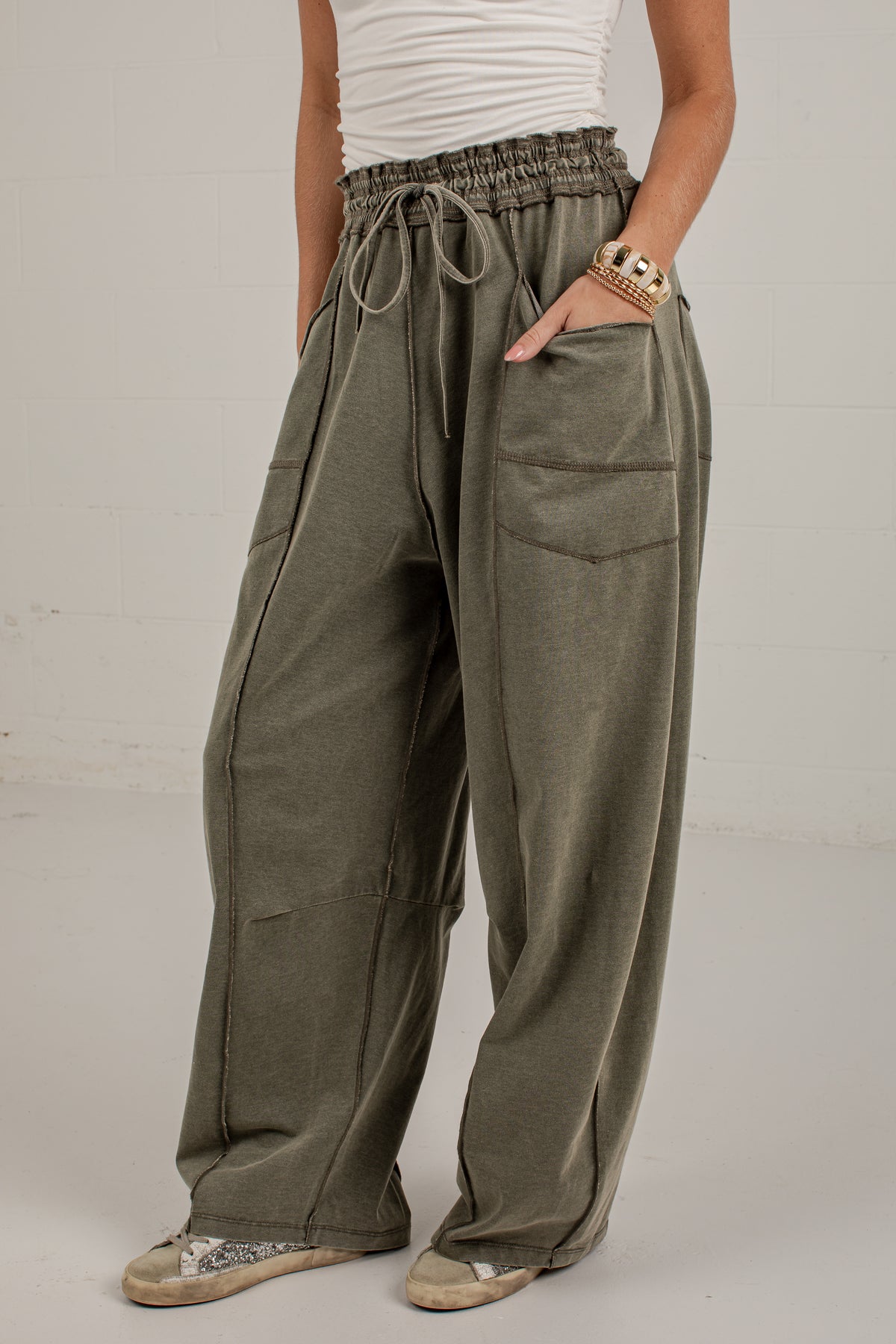 A person wearing loose, wide-leg olive green drawstring pants with large front patch pockets, paired with a white ribbed top and patterned shoes, standing against a light gray wall.