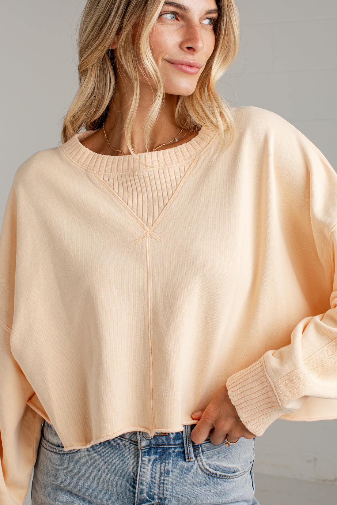 A woman wearing a light peach-colored cropped crewneck sweatshirt with a ribbed V-neck detail, paired with light-wash blue jeans.