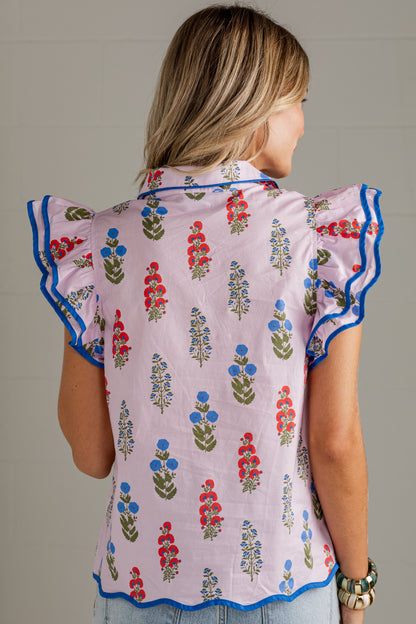 A woman shown from the back wearing a light pink short-sleeve blouse with blue ruffled trim and a colorful floral block-print pattern, paired with light blue jeans against a neutral gray background.