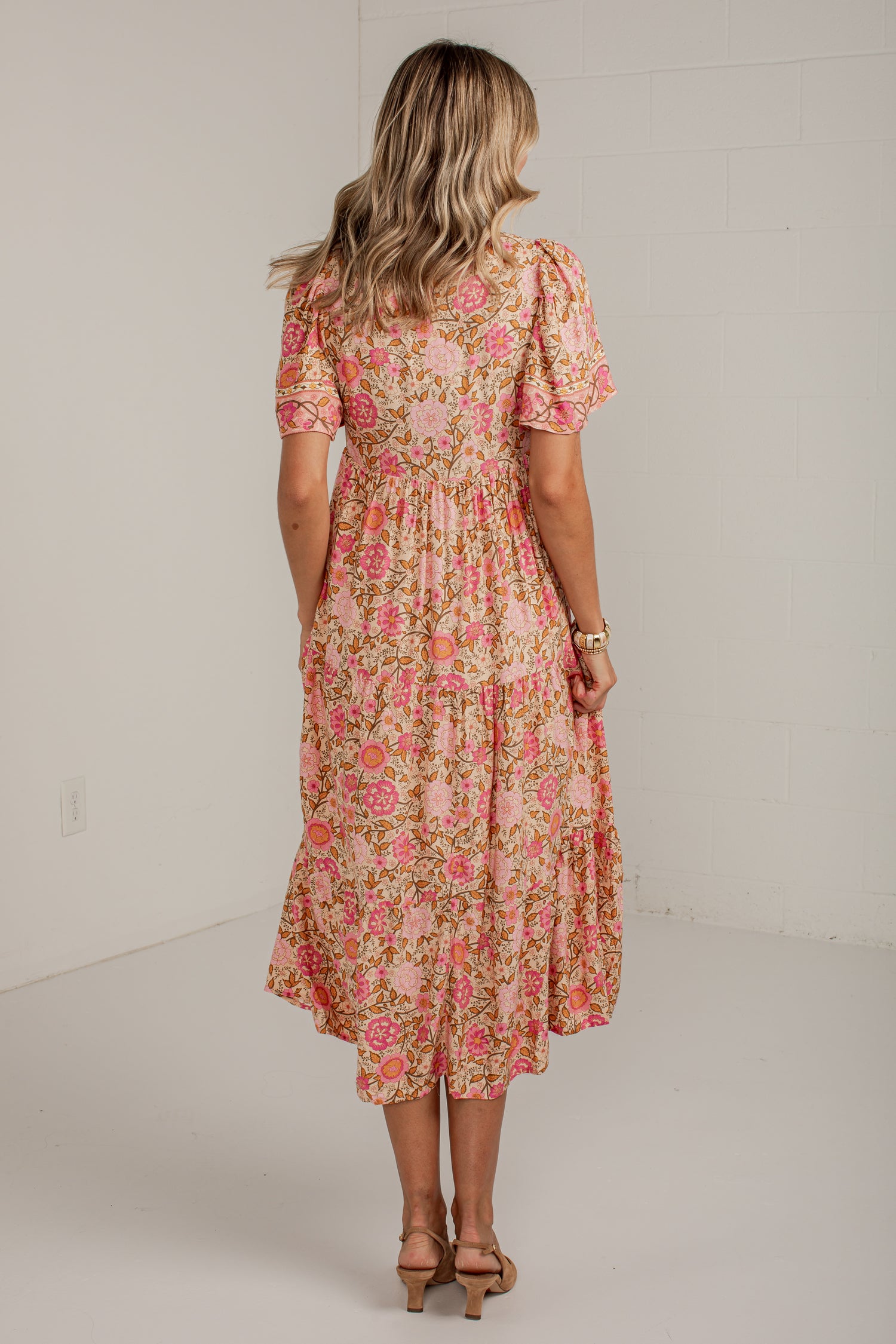 A woman viewed from behind wearing a short-sleeve midi dress with a pink and green floral pattern, standing in a bright studio with a white wall.