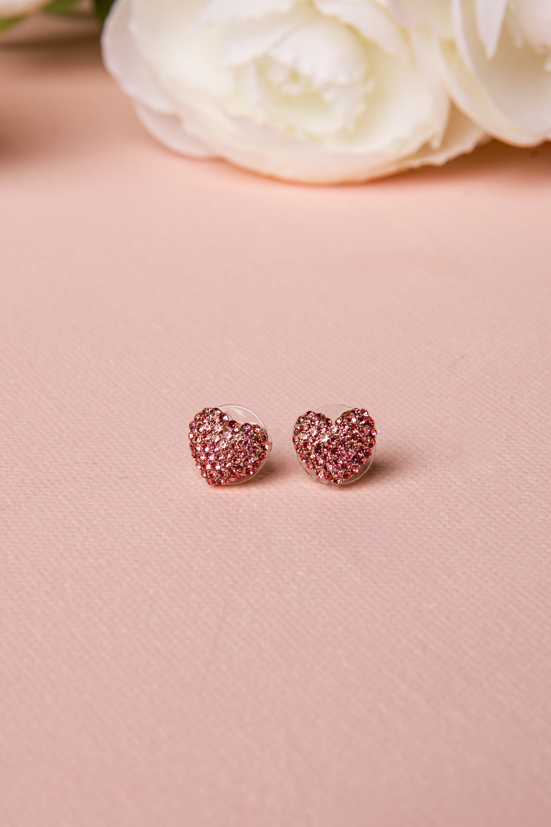 Rose gold heart-shaped earrings on a pink surface with white flowers in the background