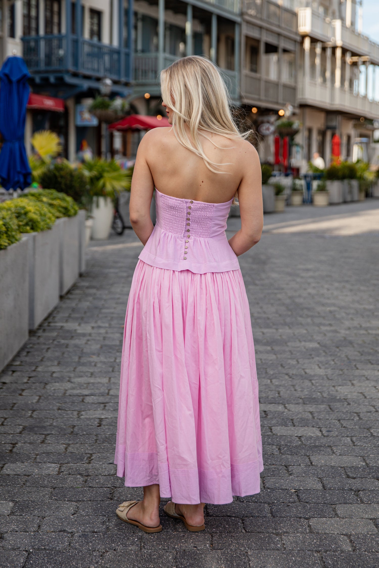 Woman in a pink dress walking on a street with shops and plants in the background