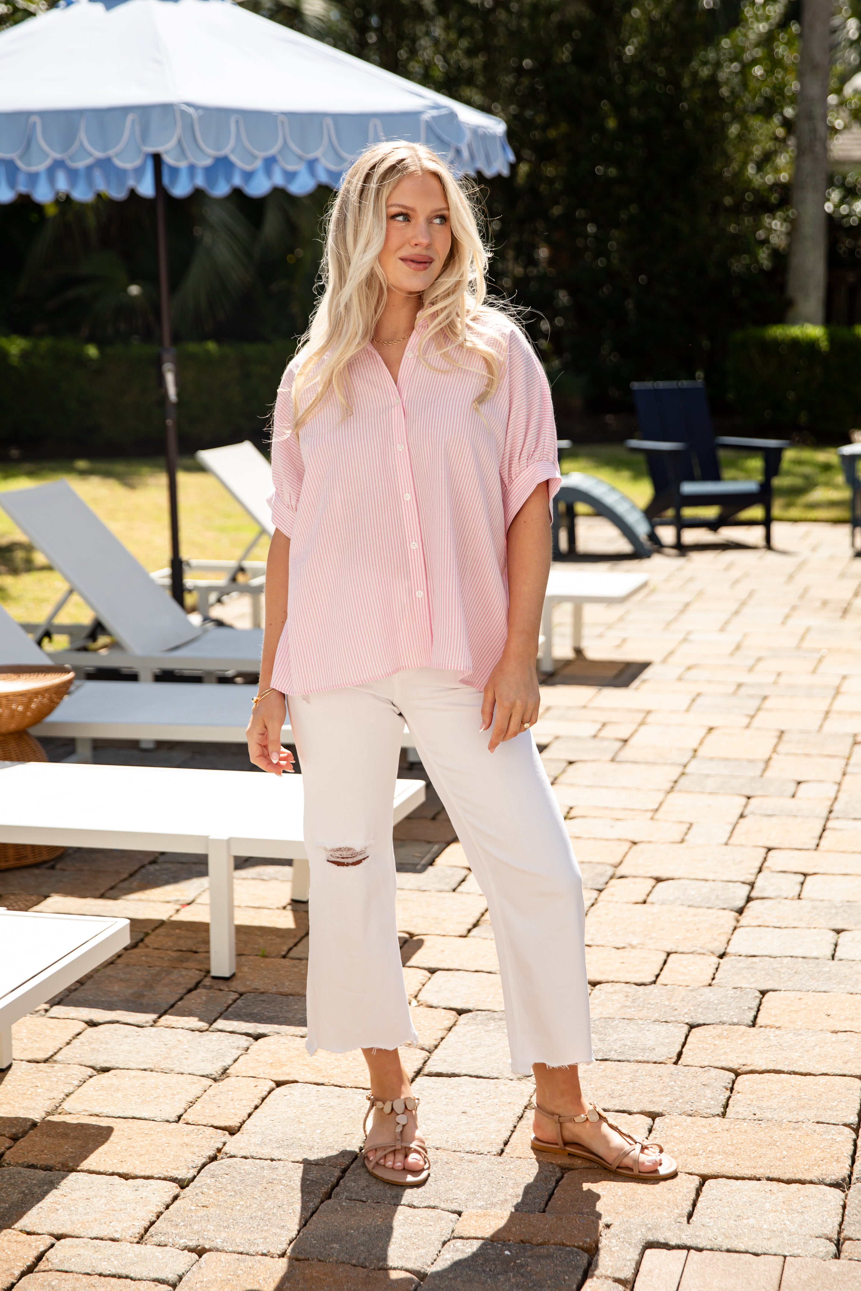 Woman in a pink shirt and white pants standing on a patio with outdoor furniture and umbrellas.