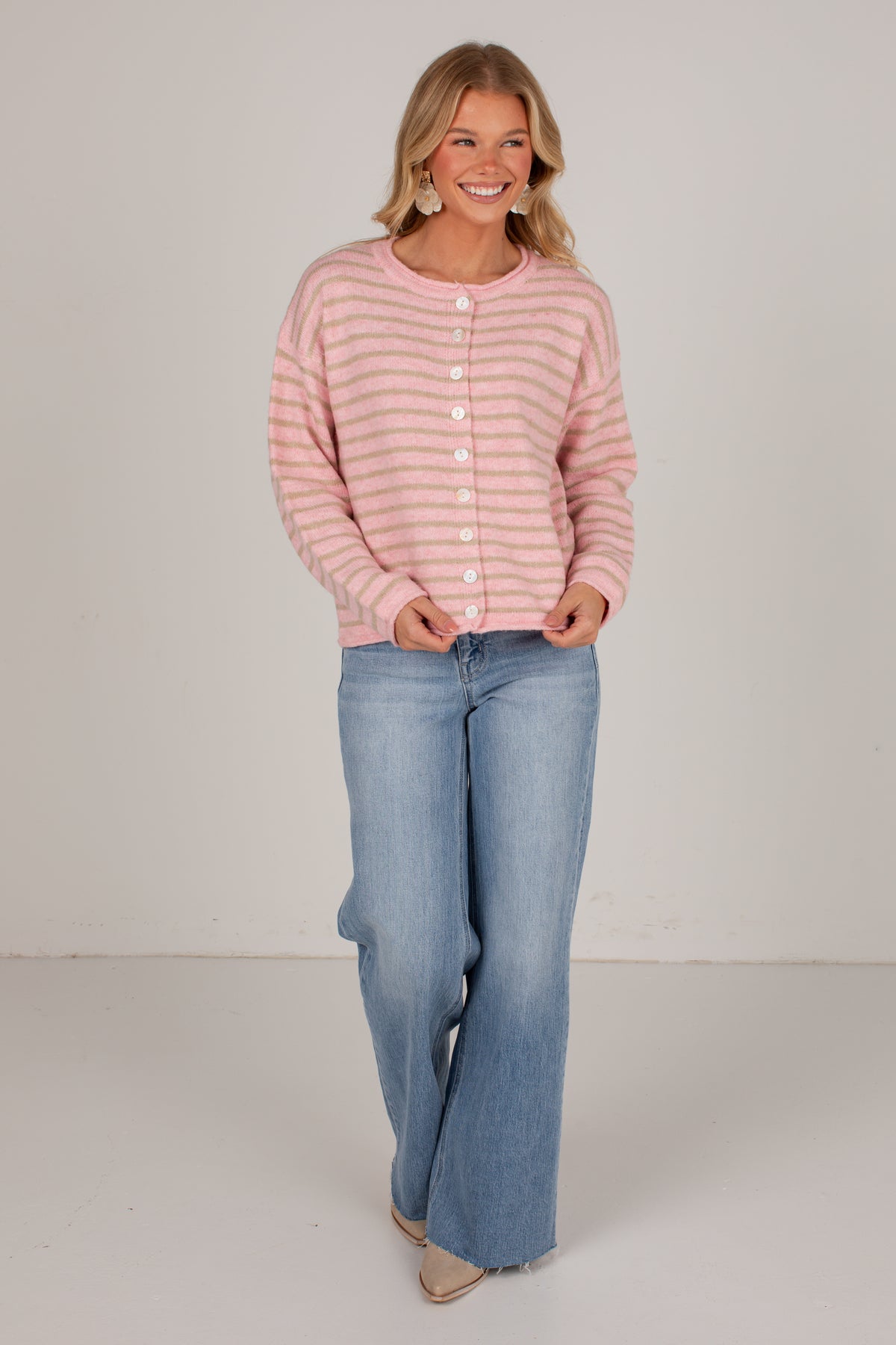 A woman modeling a pink and white striped button-up cardigan sweater with light-wash blue flare jeans, standing against a plain light gray studio background.