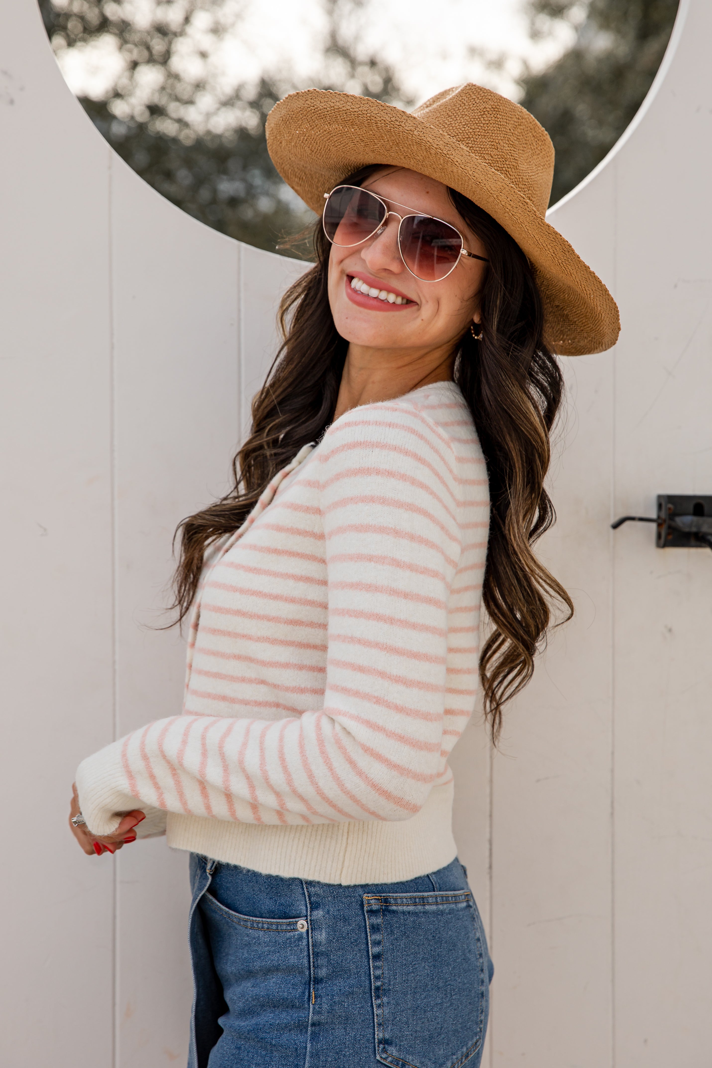Woman wearing a striped sweater, sunglasses, and a hat against a white wall.