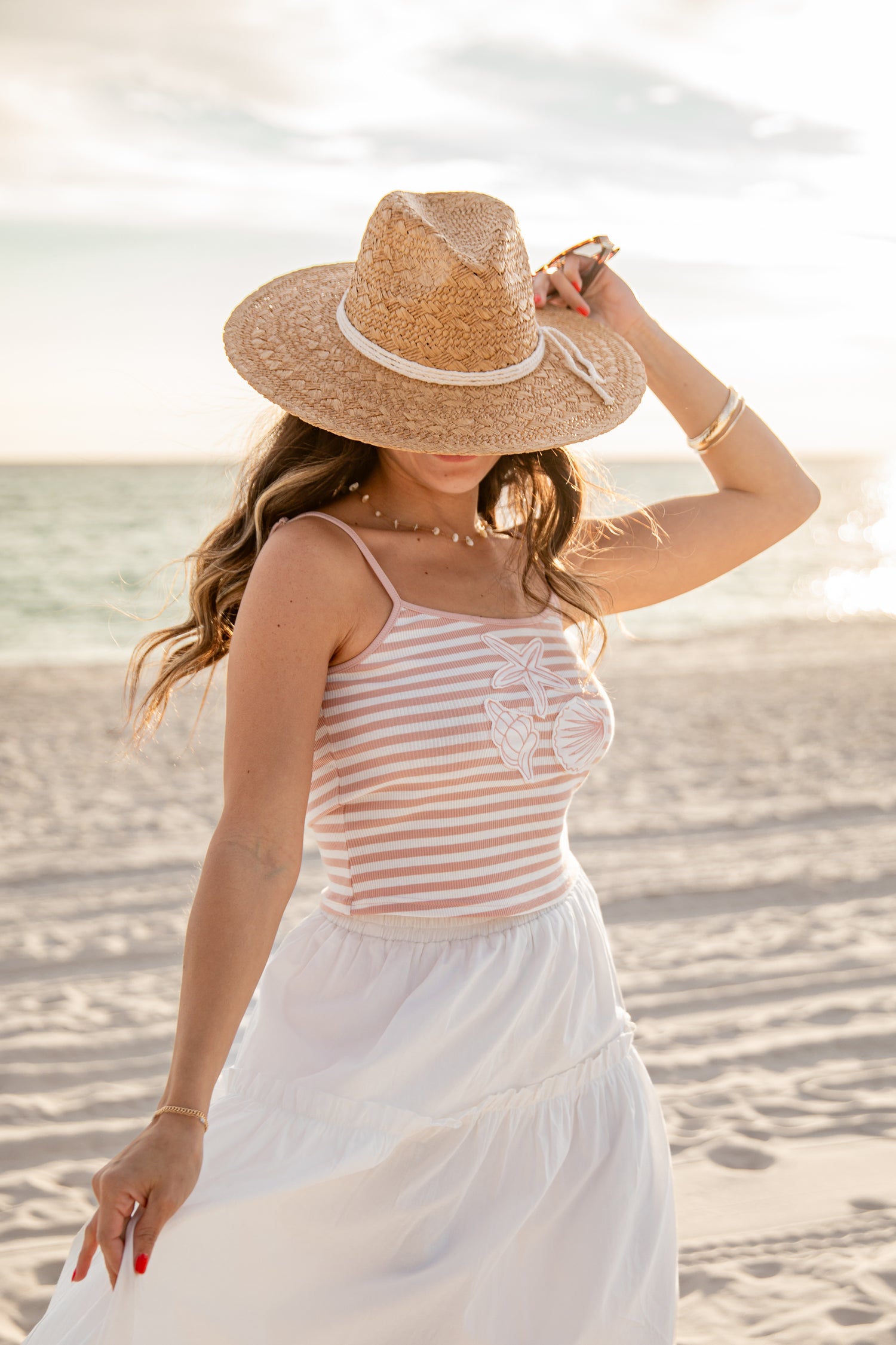 Woman in a striped tank top and white skirt with a straw hat on a beach.