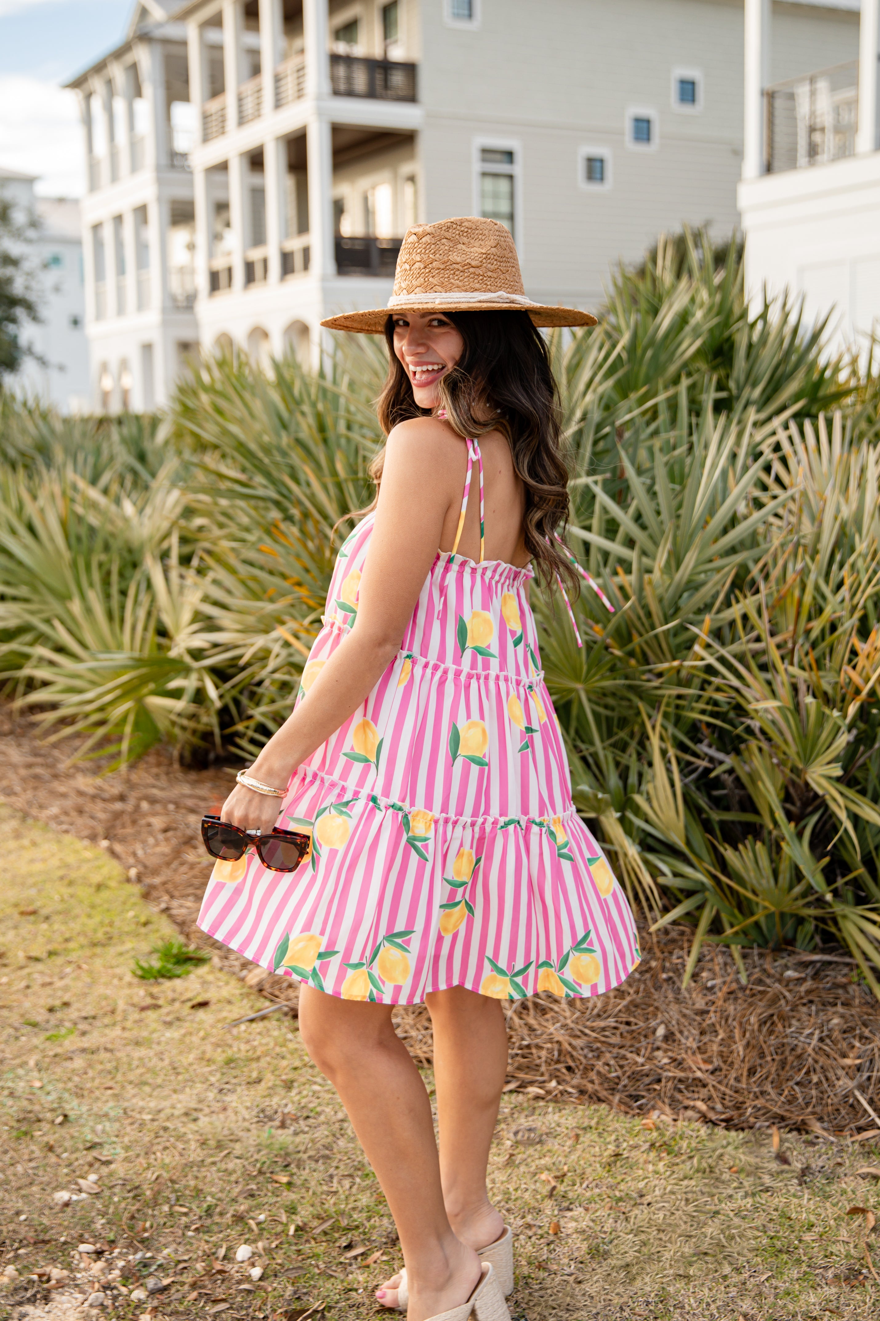 Woman in a pink dress with lemon pattern and straw hat standing outdoors.