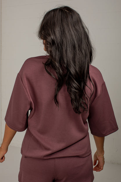 A woman viewed from behind wearing a matching mauve-brown short-sleeve top and shorts set against a plain off-white wall.