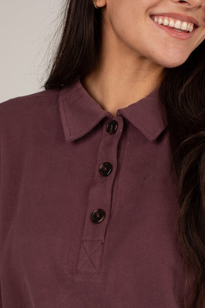 Close-up of a woman wearing a dusty plum purple polo shirt with three dark buttons and a pointed collar.