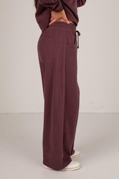 A person shown from the waist down wearing loose, wide-leg drawstring pants in a muted plum-brown color, paired with white sneakers against a plain off-white studio background.