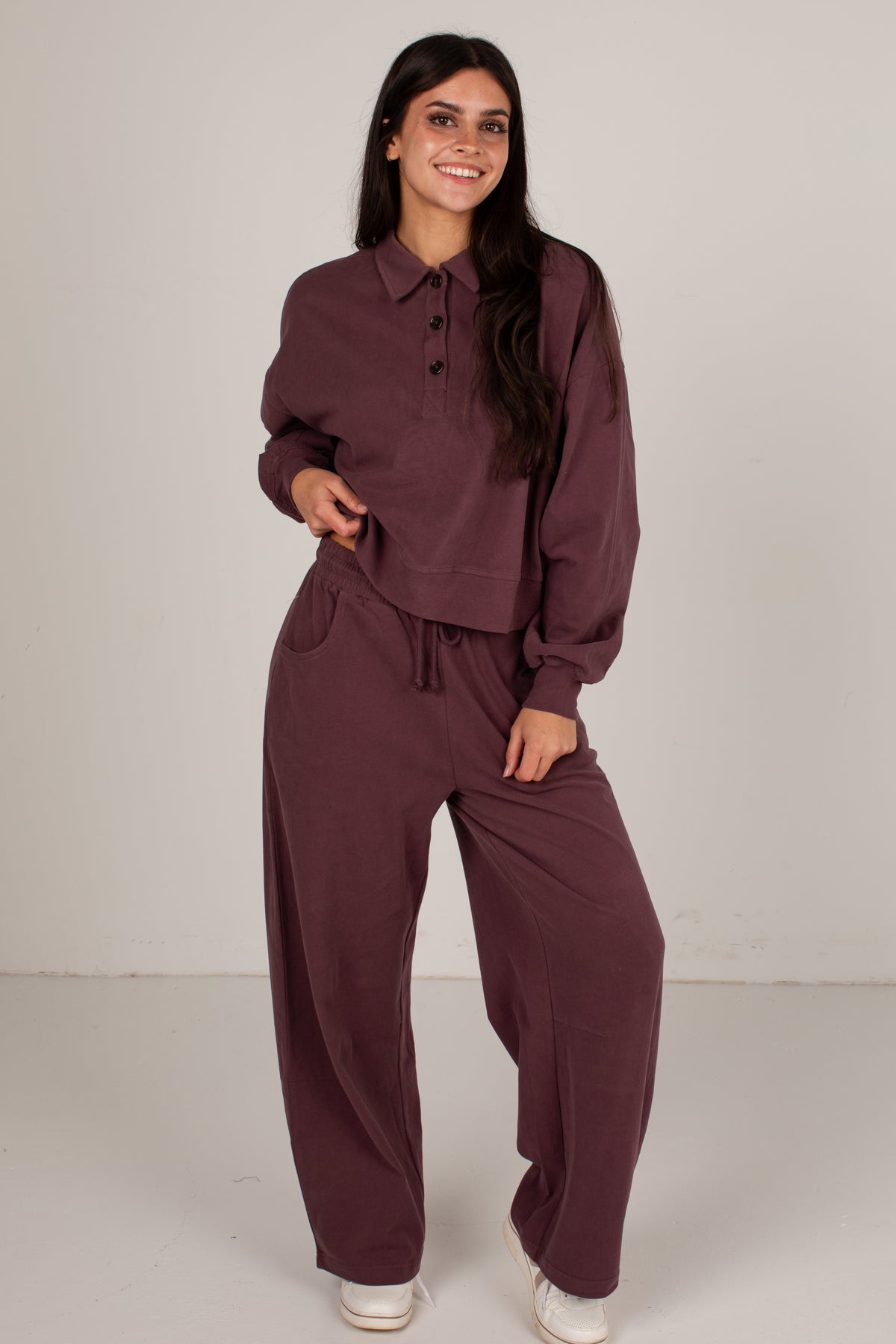 A woman modeling a matching two-piece loungewear set in a deep plum purple color, consisting of a long-sleeve button-up shirt and wide-leg drawstring pants, set against a plain light gray studio background.
