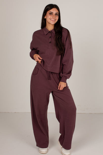 A woman modeling a matching two-piece loungewear set in a deep plum purple color, consisting of a long-sleeve button-up shirt and wide-leg drawstring pants, set against a plain light gray studio background.